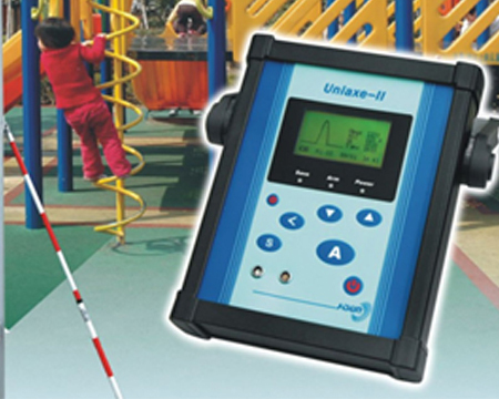 The image shows a handheld device with a digital display and various buttons, designed for testing and analysis. The background features a playground with a child climbing a play structure, and a white cane is visible, suggesting accessibility considerations.