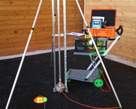 The image features a surveying setup with a tripod holding measurement equipment. There is an orange tool box, a cart with additional tools, and a visible marker on the ground, all set in a wooden interior space.