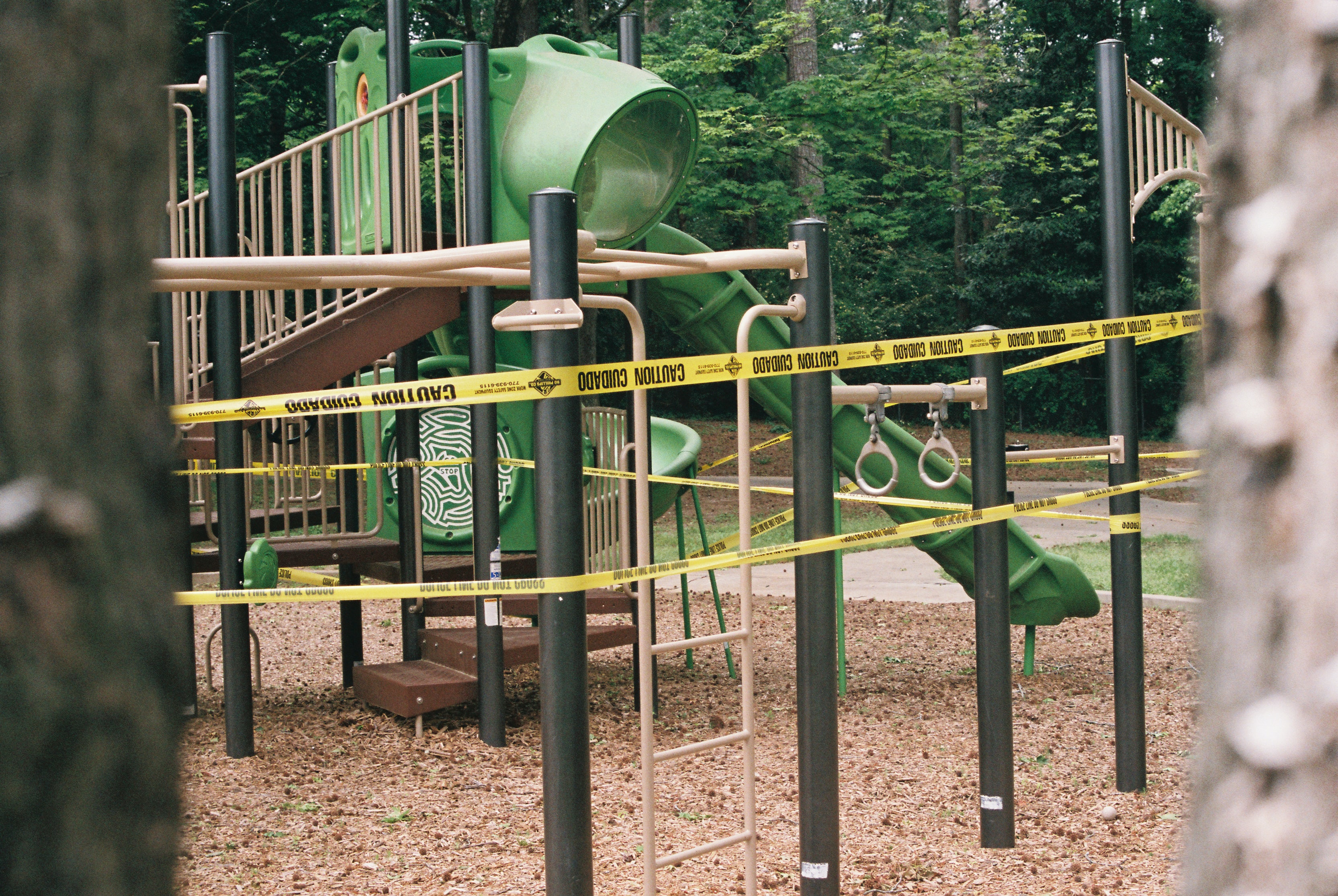 The image shows a playground structure surrounded by caution tape. The playground equipment features a green slide and climbing apparatus, with safety measures in place due to potential hazards. The area is wooded, adding a natural backdrop to the play space.