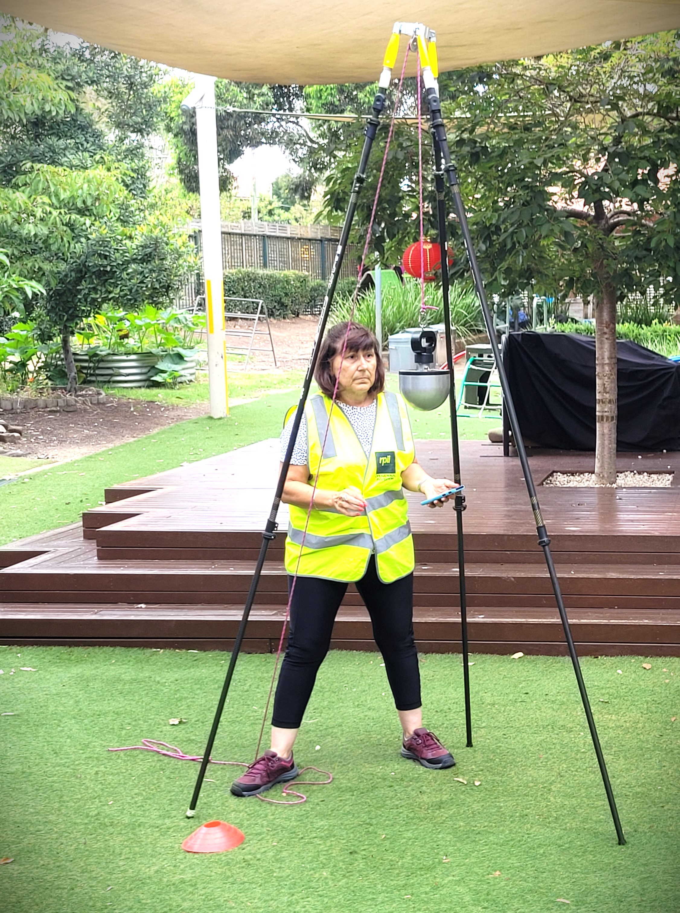 An individual wearing a high-visibility vest is standing in a park-like setting, next to a tripod structure. The person appears to be engaged in an inspection process, possibly involving equipment suspended from the tripod, with greenery and wooden decking in the background.