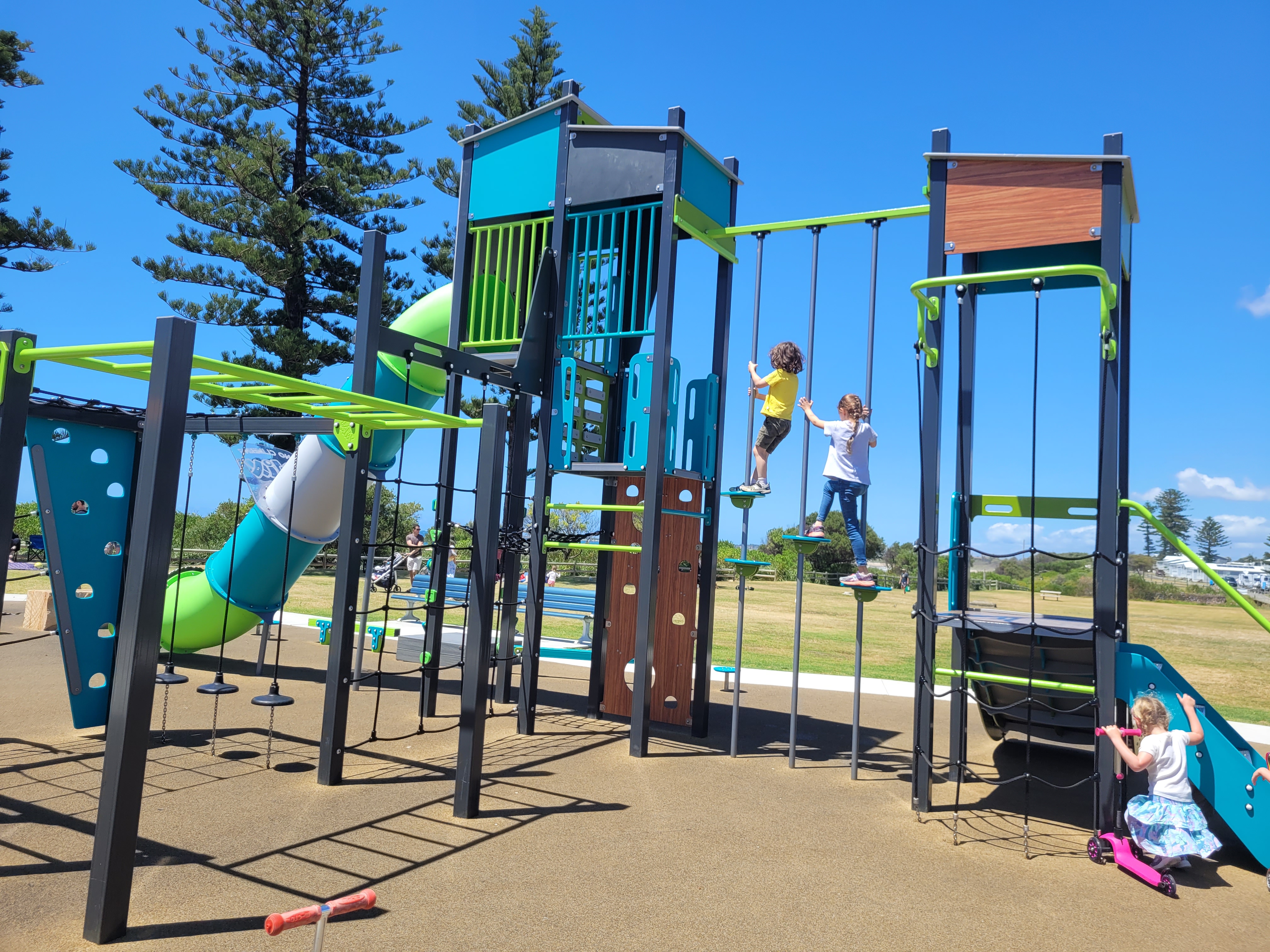 The image features a modern playground with various climbing structures and a large slide. Children are actively engaging with the equipment, climbing and playing on different sections of the playground. The area is surrounded by green grass and tall trees, with a clear blue sky above, creating an inviting outdoor space for play.