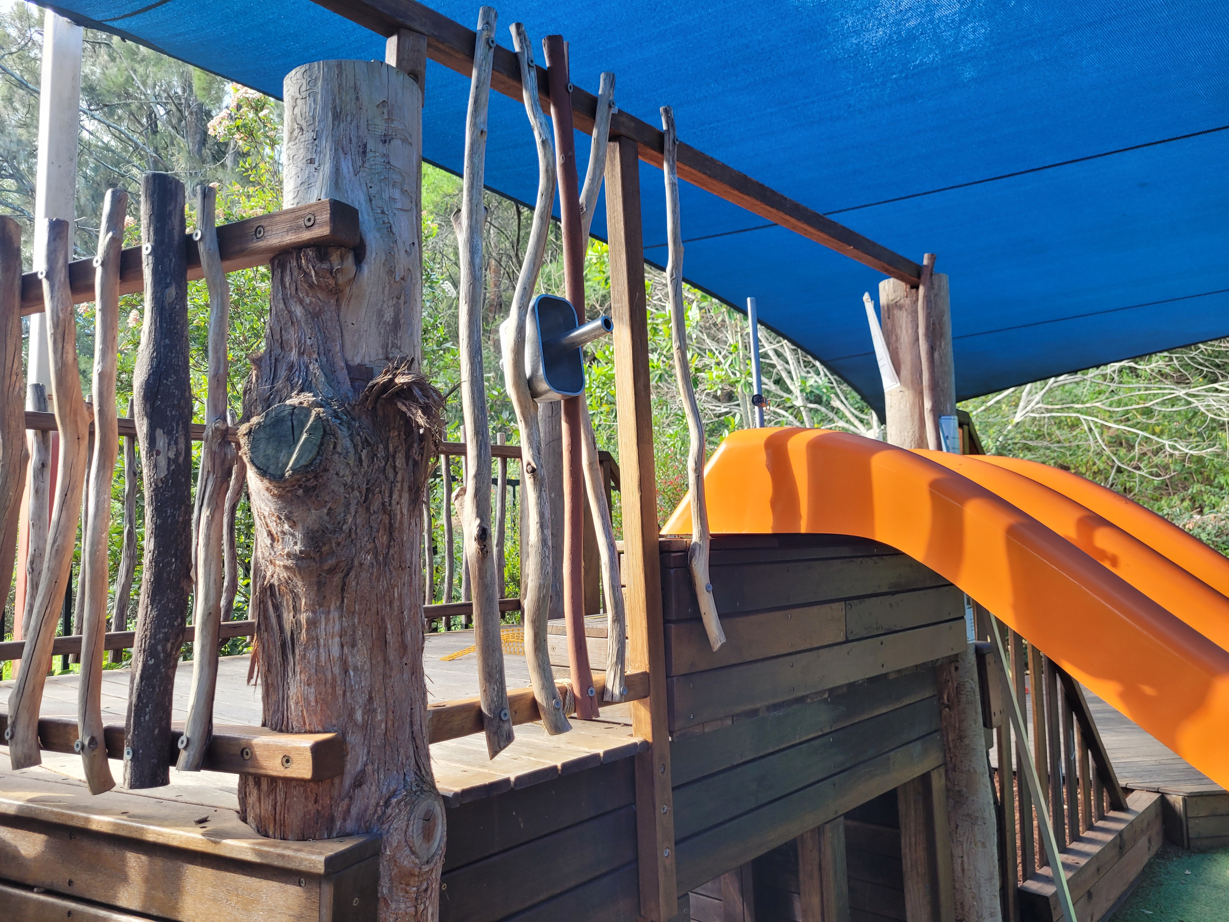 The image features a wooden playground structure with a vibrant orange slide. The play area is shaded by a blue canopy, and wooden logs create a natural barrier along the sides. In the background, greenery is visible, suggesting a natural environment.