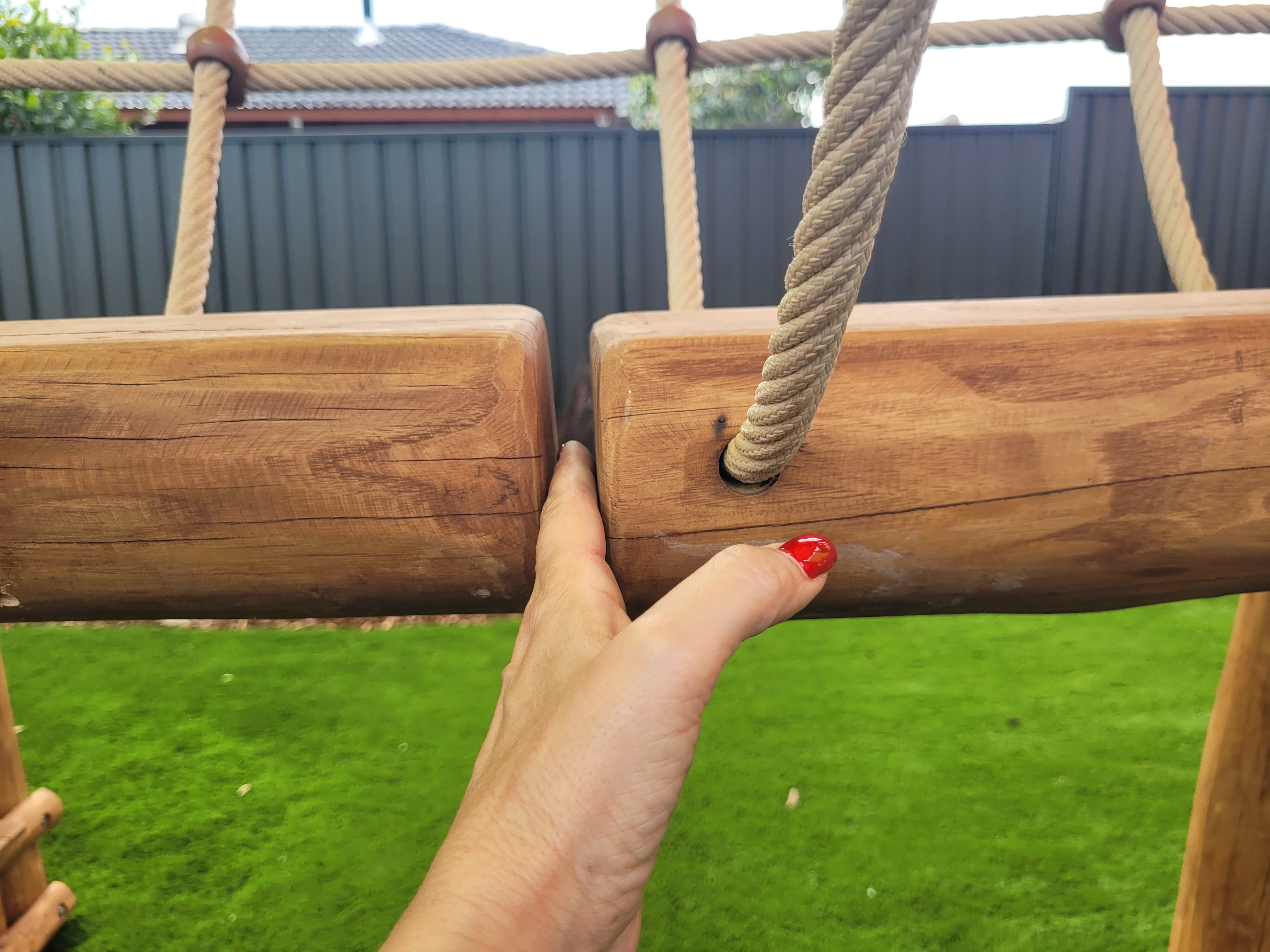 A close-up view of a wooden playground structure with two horizontal beams connected by thick ropes. A hand with red nail polish is gripping one of the beams, showcasing the natural wood finish and the texture of the material.