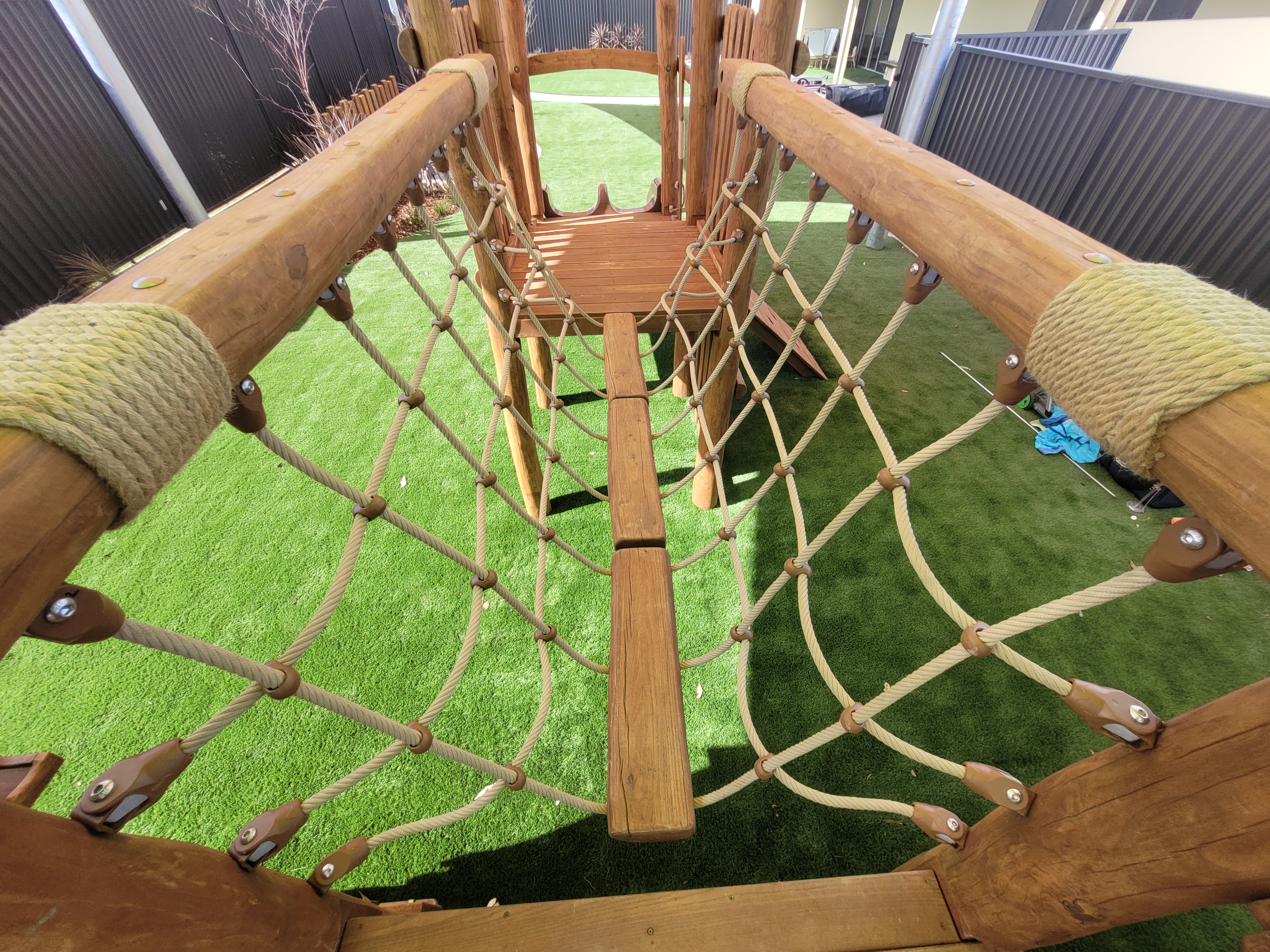 The image showcases a wooden playground structure featuring a climbing net made of ropes and wooden beams. The ground is covered with artificial grass, providing a soft landing area. Sunlight casts shadows on the equipment, highlighting the robust construction and outdoor setting.