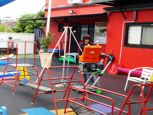 The image shows a playground area featuring colourful rubber flooring and various equipment such as a swing set and climbing structures. A woman is pushing a cart with an orange toolbox, indicating maintenance or inspection activity. The backdrop is a vibrant red wall with large windows, enhancing the lively atmosphere of the space. There are some benches and additional play equipment in the area.